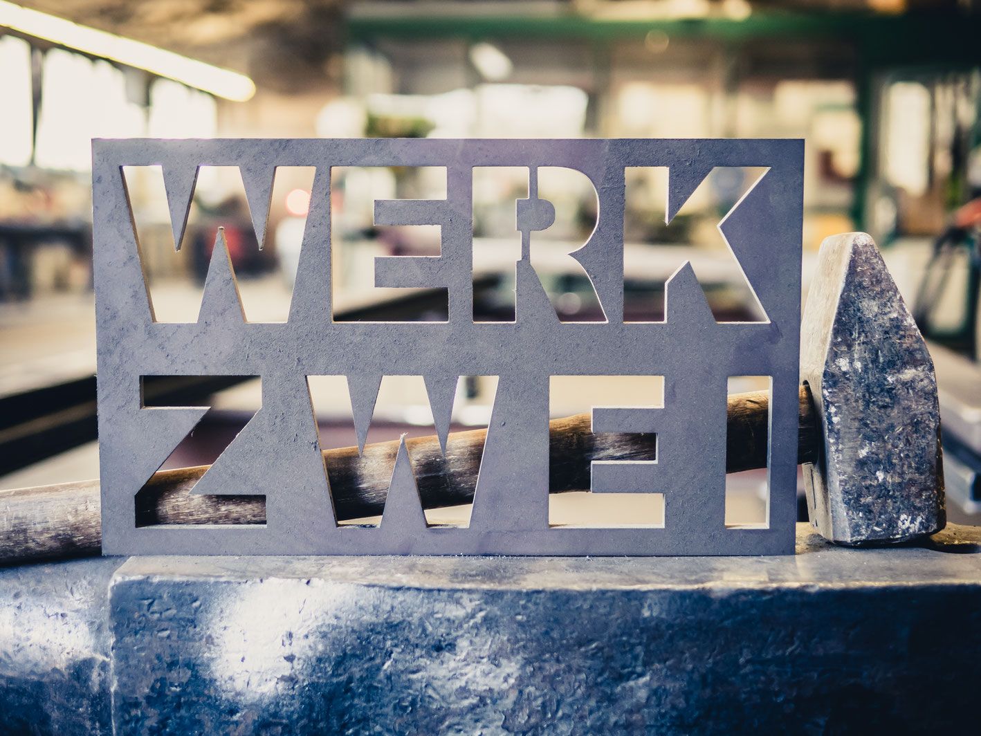 Firmenschild WERK ZWEI aus Stahlblech auf einem Amboss, daneben ein Hammerkopf.