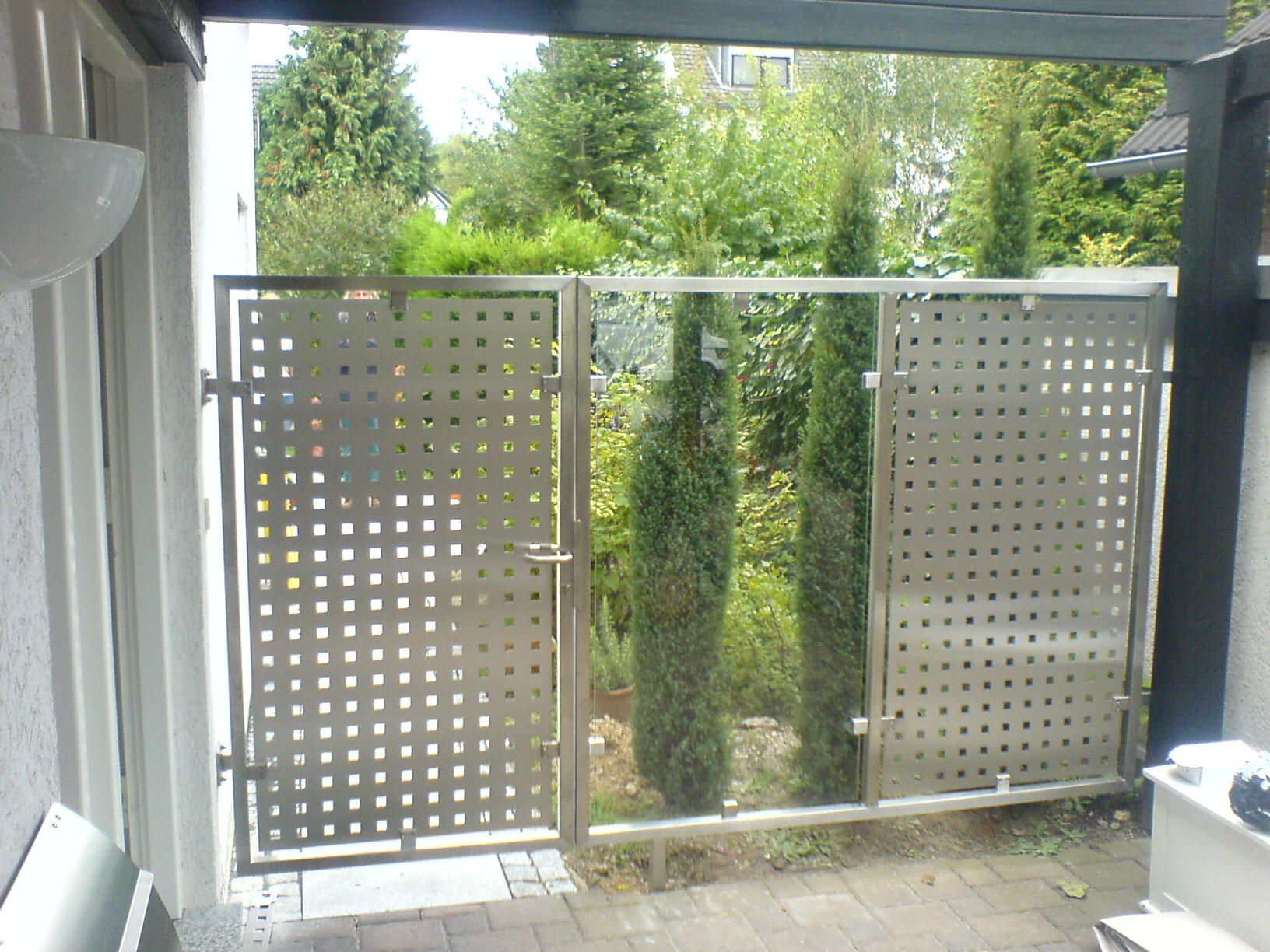 Gartentür mit Füllung aus Lochblech zwischen zwei festen Feldern im Edelstahlrahmen, mit Blick in den Garten.