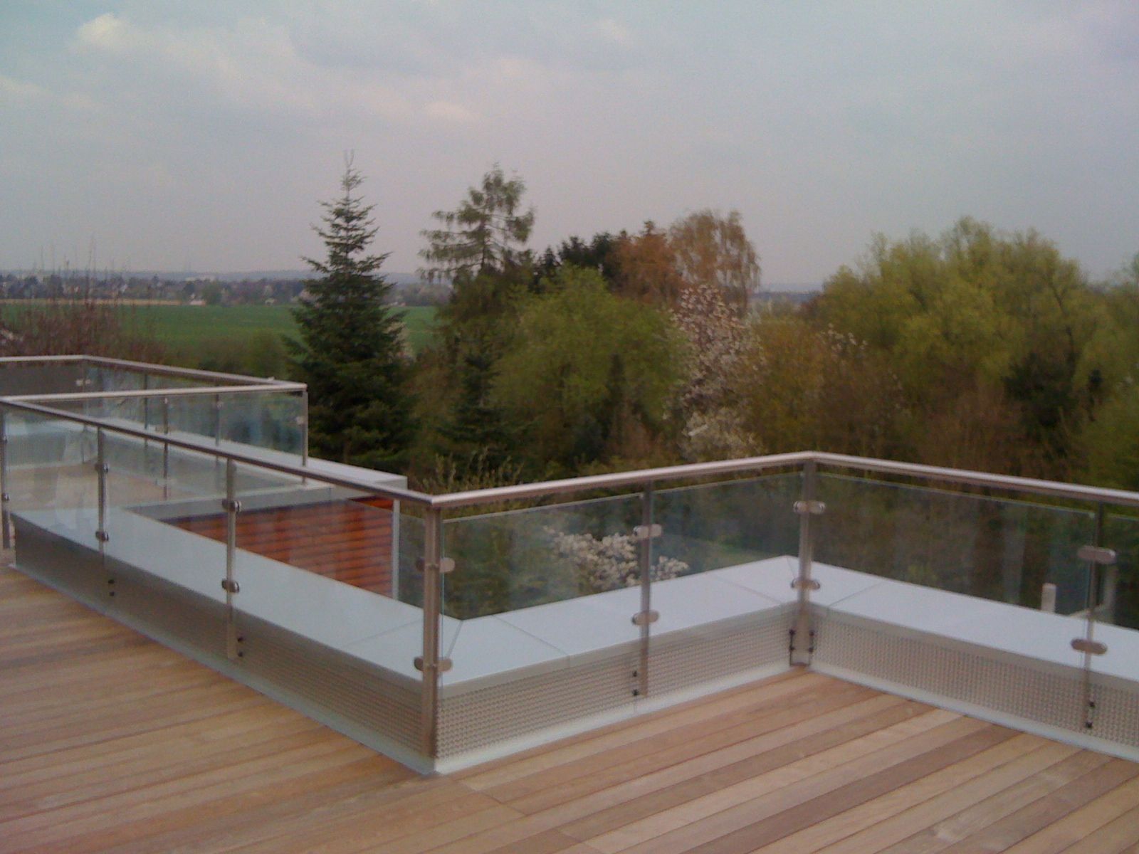 Glasgeländer mit Edelstahlpfosten und Edelstahl-Handlauf auf einer Dachterrasse mit Holzdeck und Blick ins Land.