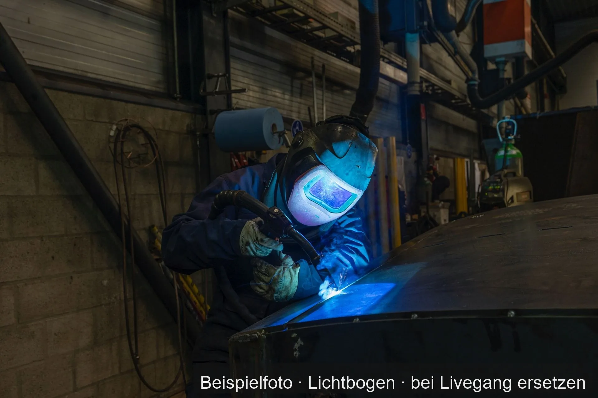 Ein Schweißer mit Schweißhelm und Lederhandschuhen setzt in einer Werkhalle eine Naht an einer großen Stahlblechhaube.